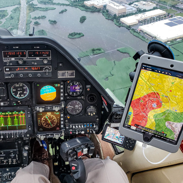 View from a cockpit with a pilot's hands and legs visible. The control panel displays various instruments and gauges. An electronic tablet, mounted to the side, shows a colorful map indicating weather conditions. Below, a landscape with buildings and water is visible.