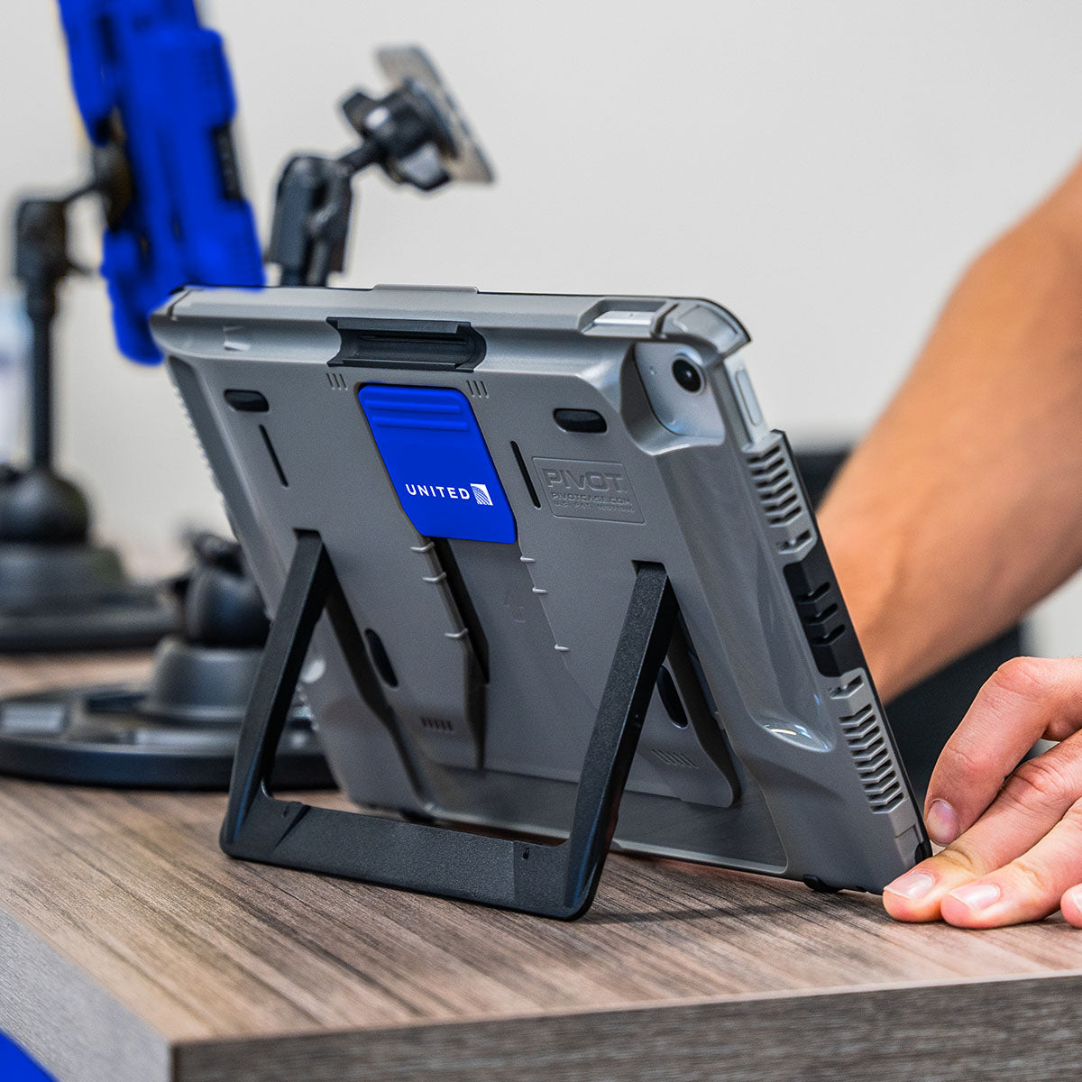 A person using a tablet in a rugged case with a built-in stand on a wooden desk. The tablet has a blue clip labeled "United." In the background, there is another device with a blue holder attached to a mount.