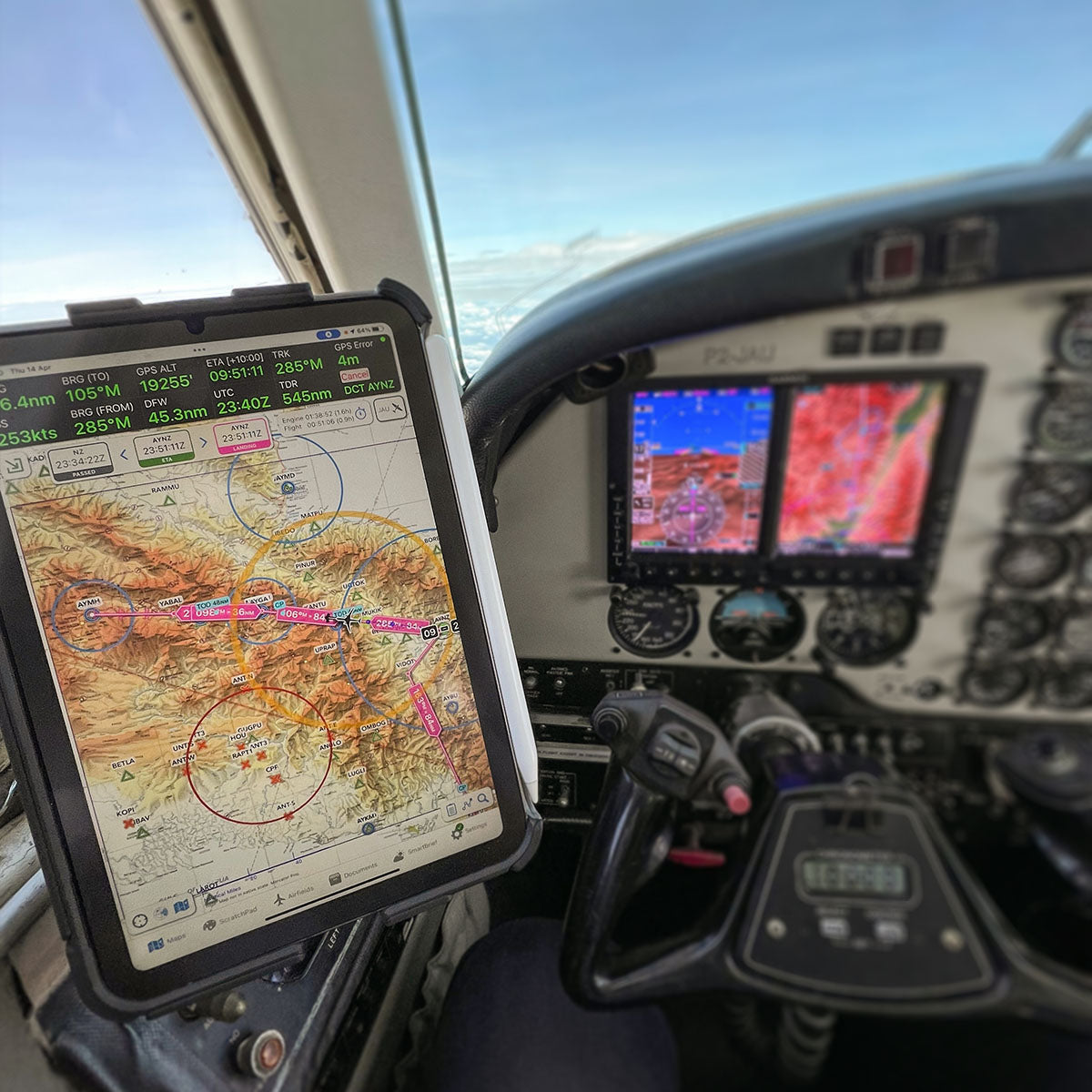 A cockpit view of an aircraft showing detailed navigation maps on a tablet fixed to the side and flight instruments displayed on screens. The scenery outside the windows is a clear sky with expansive landscapes below. Various flight controls are visible.
