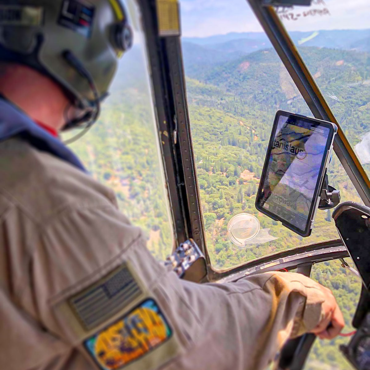 A person, wearing a helmet and a uniform with an American flag patch, pilots a helicopter. They are looking at a tablet mounted within the cockpit. The view outside shows a lush, mountainous landscape under a clear sky.