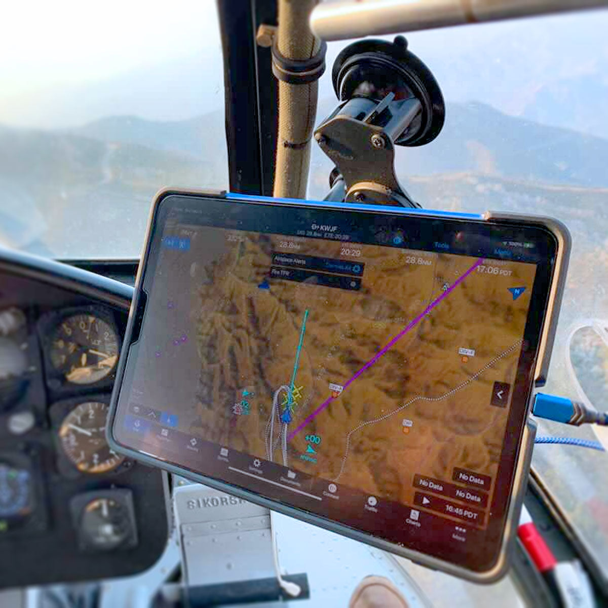An electronic flight navigation system is displayed on a tablet mounted within an aircraft cockpit. The screen shows a flight path over a mountainous terrain, including various waypoints and data points. Aircraft instruments and a partial view out the window are also visible.