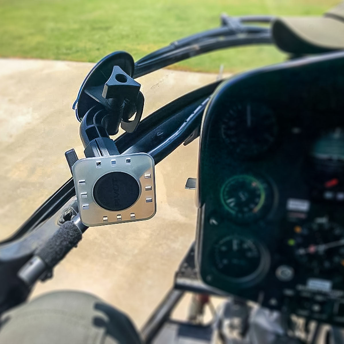 Close-up view of the cockpit of a helicopter, focusing on a mounted electronic device on the control panel. The background shows a grassy outdoor area through the cockpit window, suggesting the helicopter is grounded.