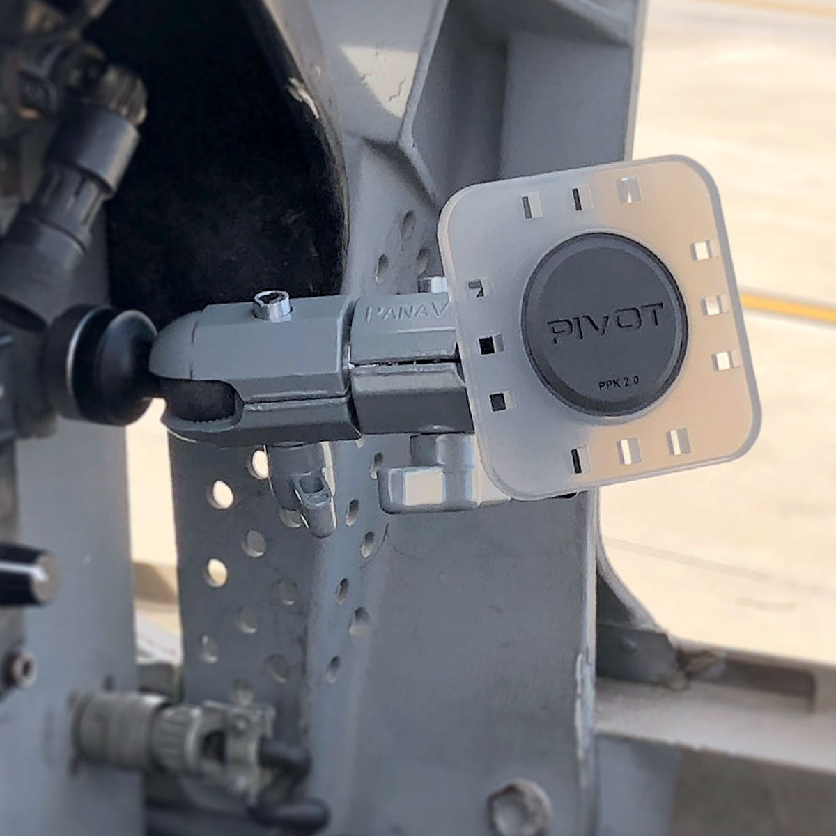 A PIVOT PPK 2.0 mount is fixed to a metallic structure. The mount has a rectangular base with rounded corners and several slots around its edge. It is attached to a swivel joint, allowing for adjustable angles. The background shows part of an industrial setting.