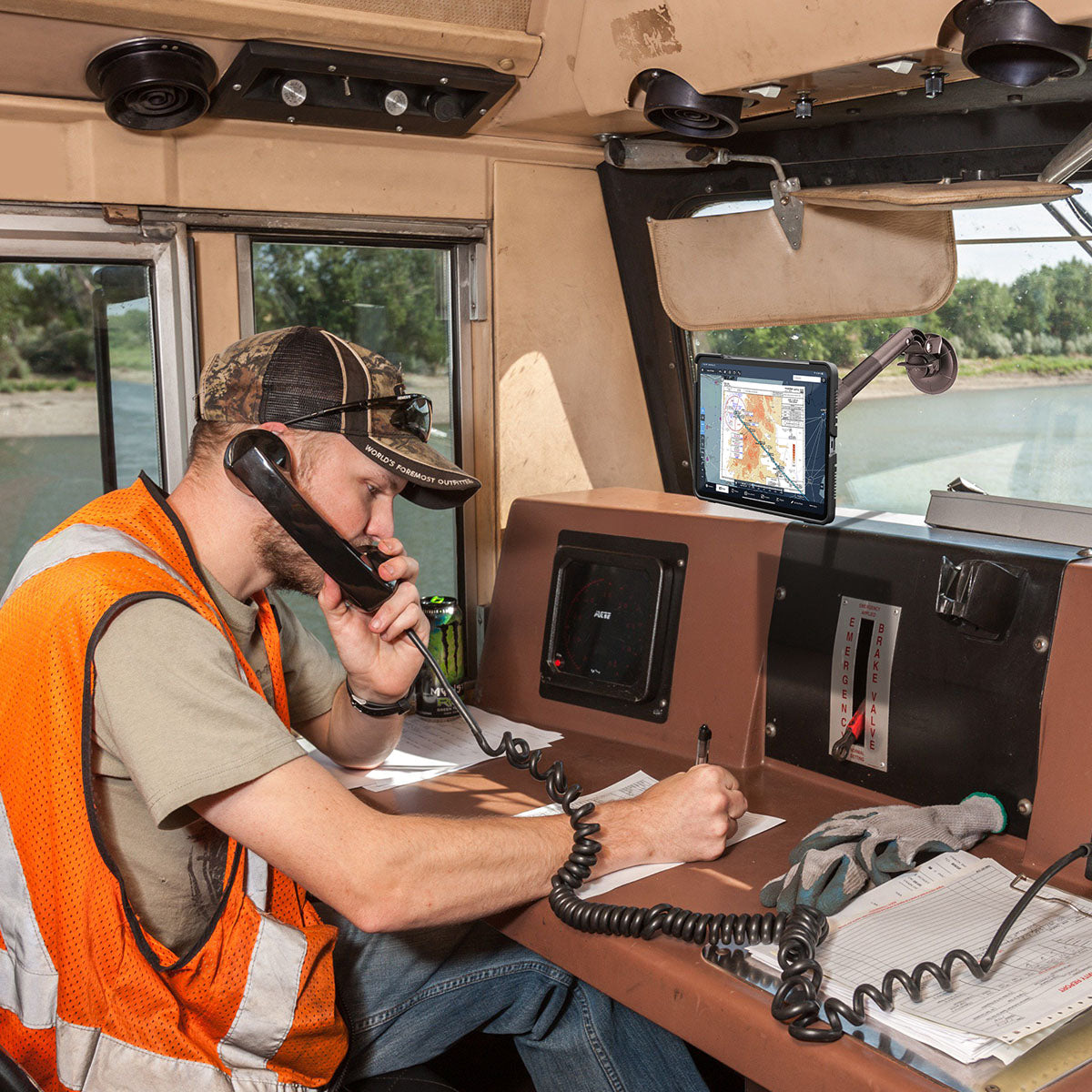A man in an orange safety vest and camouflage cap is speaking on a landline phone while writing notes. He is seated at a desk with papers, a drink can, and dual screens in front of him in what appears to be a train or vehicle cabin with windows showing outdoor scenery.