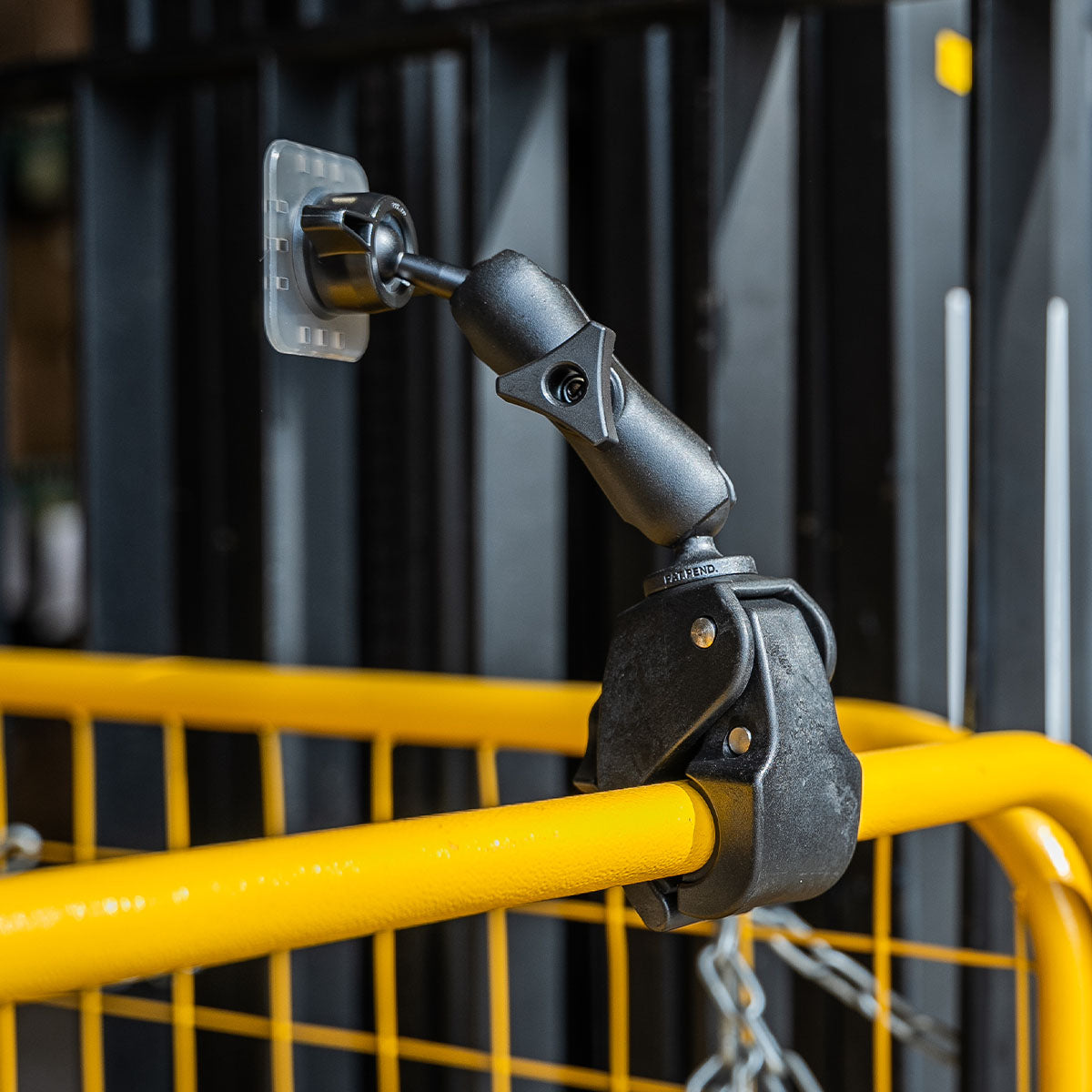 A black, adjustable mounting clamp is securely attached to a yellow metal railing. The clamp has a ball joint for versatile positioning and is pictured against a background of vertical black structures and a chain hanging partially in the view.