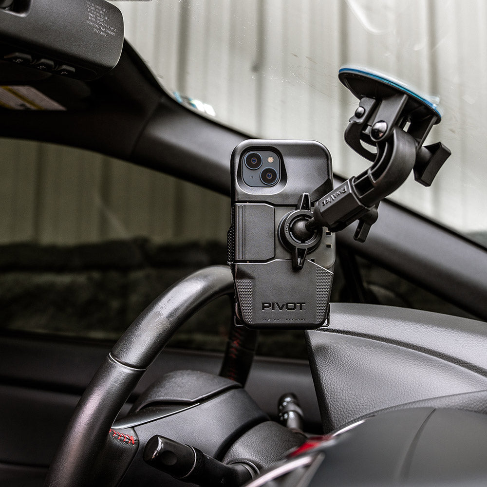 A smartphone mounted on a car's dashboard using a PIVOT M23A phone holder. The holder is attached to the windshield and has a flexible arm for adjustable positioning. The phone screen is facing the driver, and the car's steering wheel and interior are visible. Note: Does not fit Pro versions of iPhone 13 or iPhone 14.