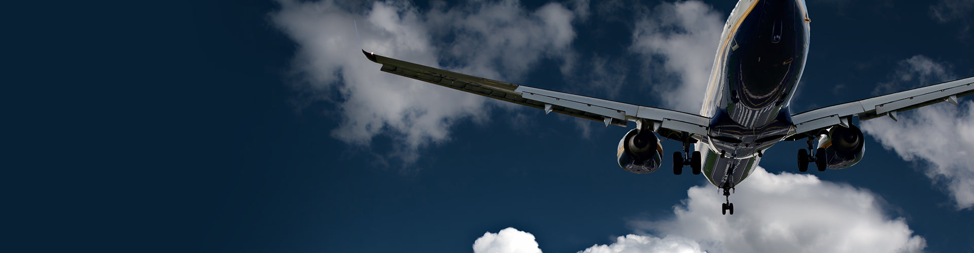 A commercial airplane with landing gear deployed descends through a cloudy sky, preparing for landing. The aircraft is viewed from below against a backdrop of a deep blue sky with white clouds, highlighting the underside and wings of the plane.