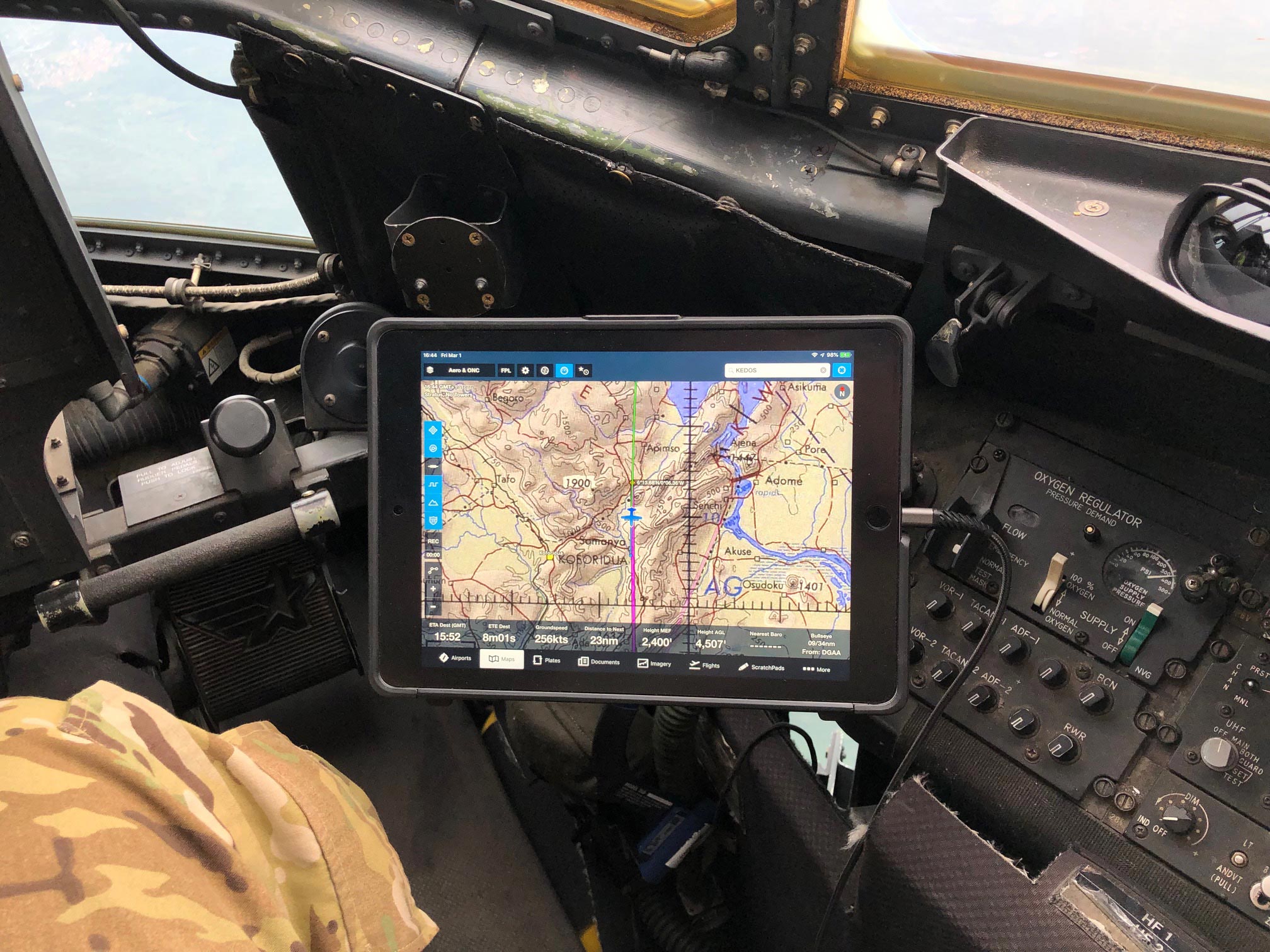 A tablet displaying a flight navigation map is mounted in the cockpit of an aircraft. The cockpit features controls, dials, and a pilot's seat. The pilot's camouflage uniform is partially visible on the left.