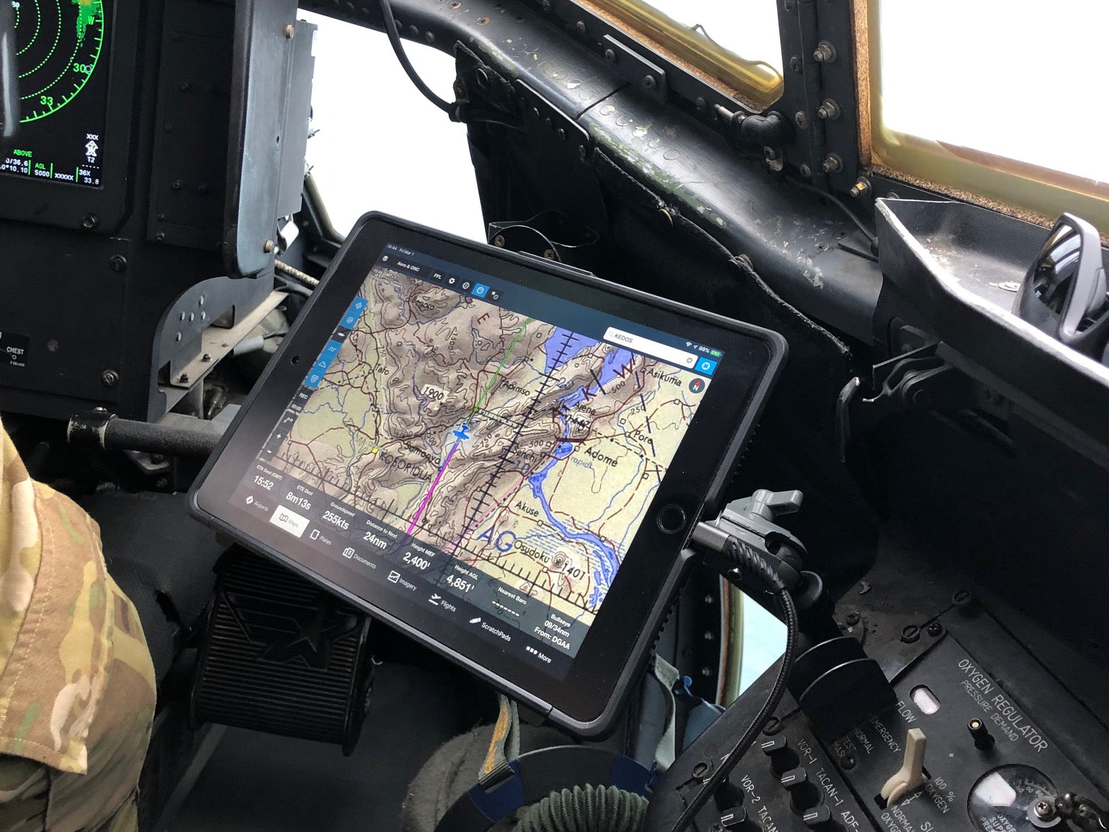 A tablet displaying a detailed map is mounted in the cockpit of an aircraft. The cockpit panel is visible, showcasing various controls and buttons. The map on the tablet shows terrain features, roads, and navigation information. A person in camouflage attire is partially visible.