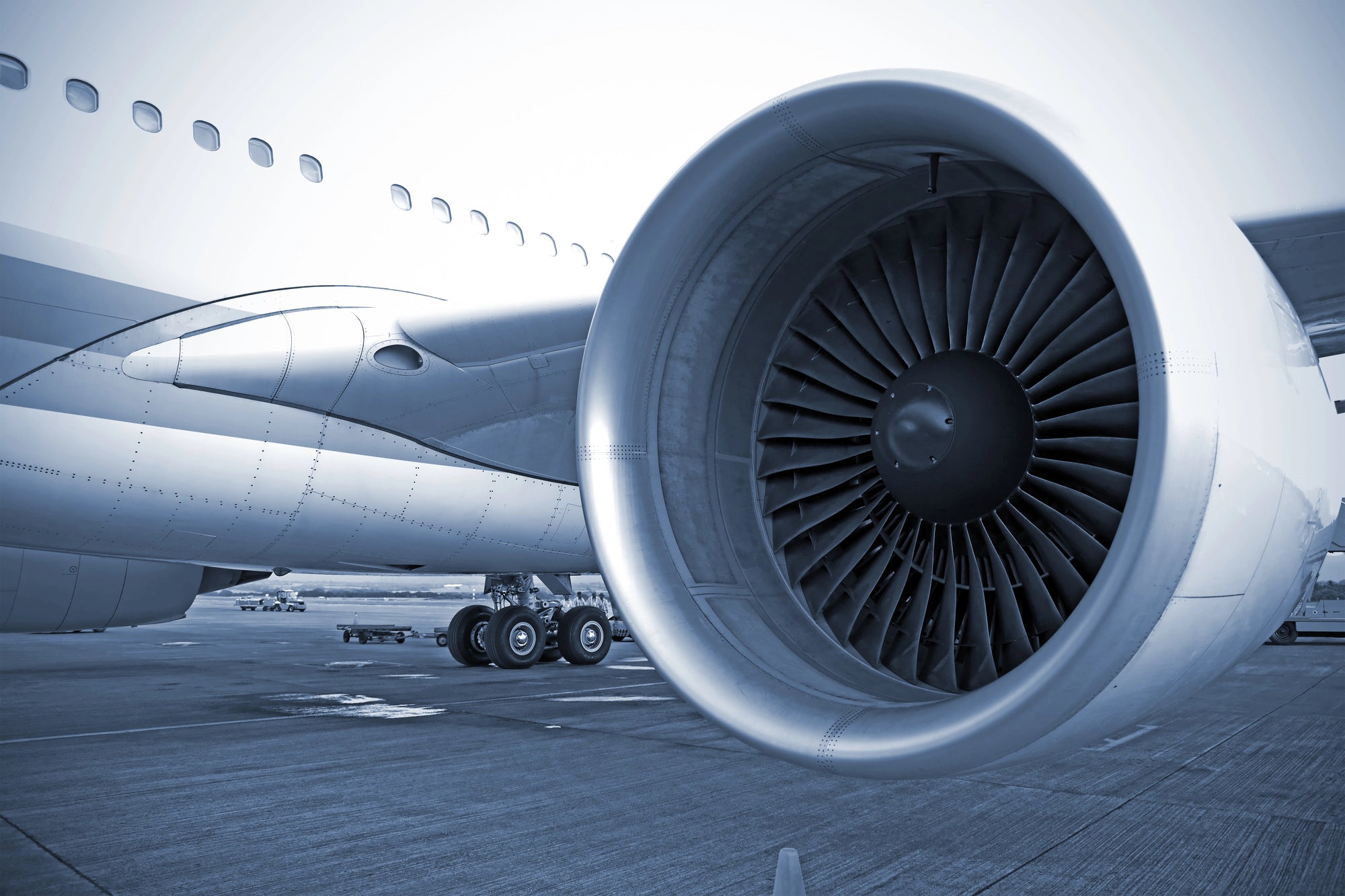 Close-up view of a large jet engine attached to the wing of a commercial aircraft. The turbine blades are prominently visible. The airplane rests on the tarmac, and the landing gear and parts of the fuselage are seen in the background.