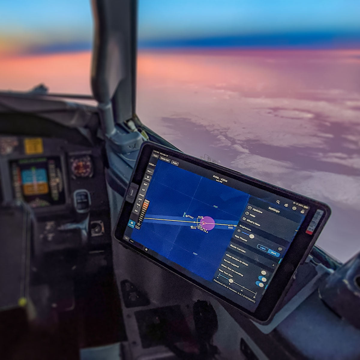 A view from inside an airplane cockpit, showing a digital flight map on a large screen. The map displays the flight route and current location. Outside the cockpit window, there's a scenic sunset sky with soft, colorful hues blending together.