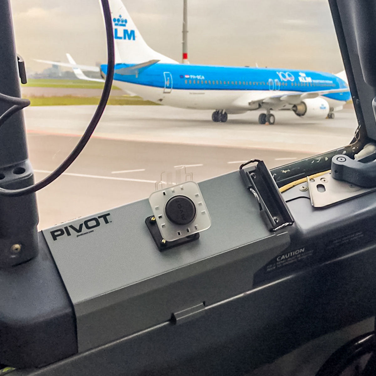 View from an aircraft cockpit, showing a KLM airplane taxiing on the runway. The cockpit has a device labeled "PIVOT" attached to a panel. The scene captures the airport tarmac and overcast sky in the background.