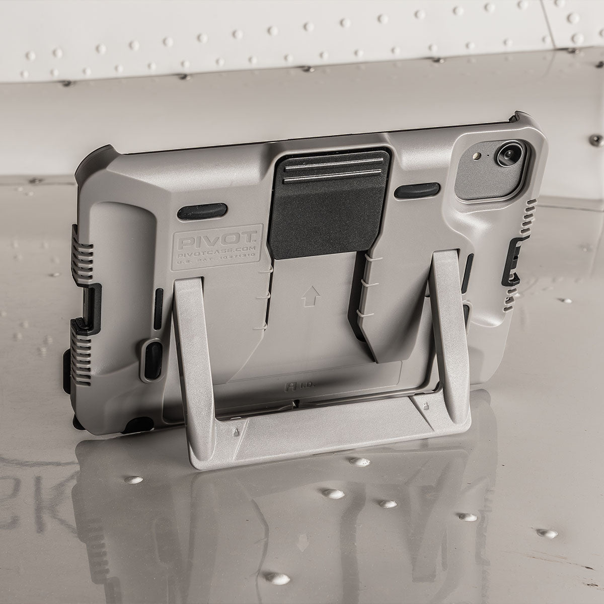 A silver tablet encased in a rugged, protective shell with a built-in kickstand is positioned on a reflective surface. The tablet is angled rear-view, showcasing its sturdy back design and the extended kickstand for hands-free use.