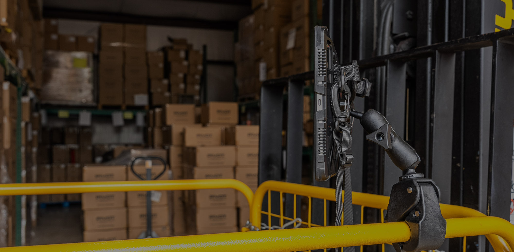 A warehouse interior with stacks of brown cardboard boxes on shelves. In the foreground, there is a yellow metal barrier and a mounted device attached to it, possibly for scanning or monitoring inventory. The background shows more boxes and a loading area.