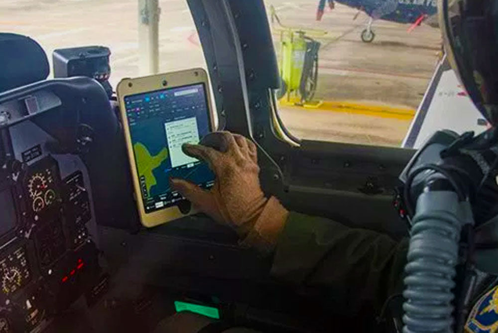 A pilot in the cockpit of an aircraft is using a touch screen tablet. The dashboard is filled with controls and instruments, and another aircraft is visible through the window at the far end of the runway. The pilot's hand is interacting with the tablet's interface.