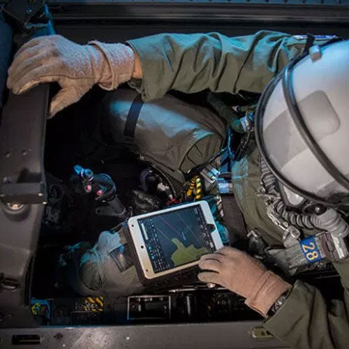 A pilot in full gear, including gloves and helmet, sits in an aircraft cockpit. The pilot is holding a tablet displaying navigational data or a map. Various controls, buttons, and instrumentation are visible around the cockpit area.