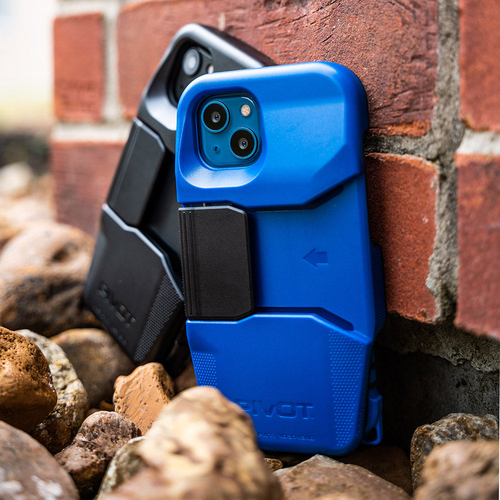 Two smartphones in rugged cases lean against a brick wall on a bed of rocks. The iPhone 13 in the foreground has a blue and black PIVOT M23A case, while the one behind it, possibly an iPhone 14, has a black PIVOT M23A case. Both cases feature an angular design and a clip attachment.