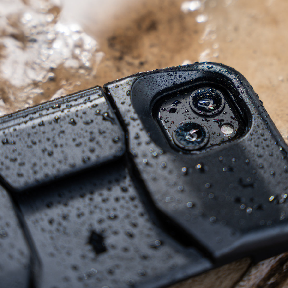 A close-up of a rugged PIVOT M23A, reminiscent of an iPhone 13 with a dual camera setup, covered in water droplets indicating water resistance. The phone is lying on a textured surface with a mix of wet areas and patches of light reflections.