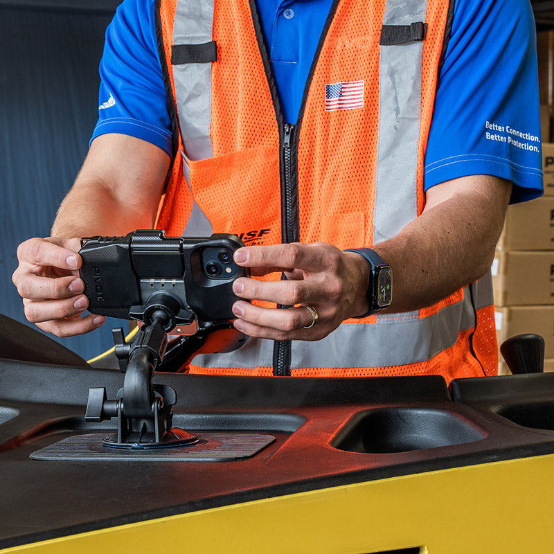 A person wearing an orange safety vest over a blue shirt uses a mounted PIVOT M23A, focusing on the screen. The vest has an American flag patch and the words “Better Connections. Better Protection.” on it.