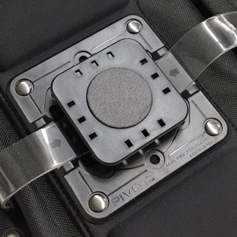 A close-up picture of a black and gray mechanical part, featuring a square base with a circular center. There are transparent straps on either side and several small rectangular slots around the center. The word "PIVOT" is visible on the base. This is the Low Profile Kneeboard Adapter by PIVOT.