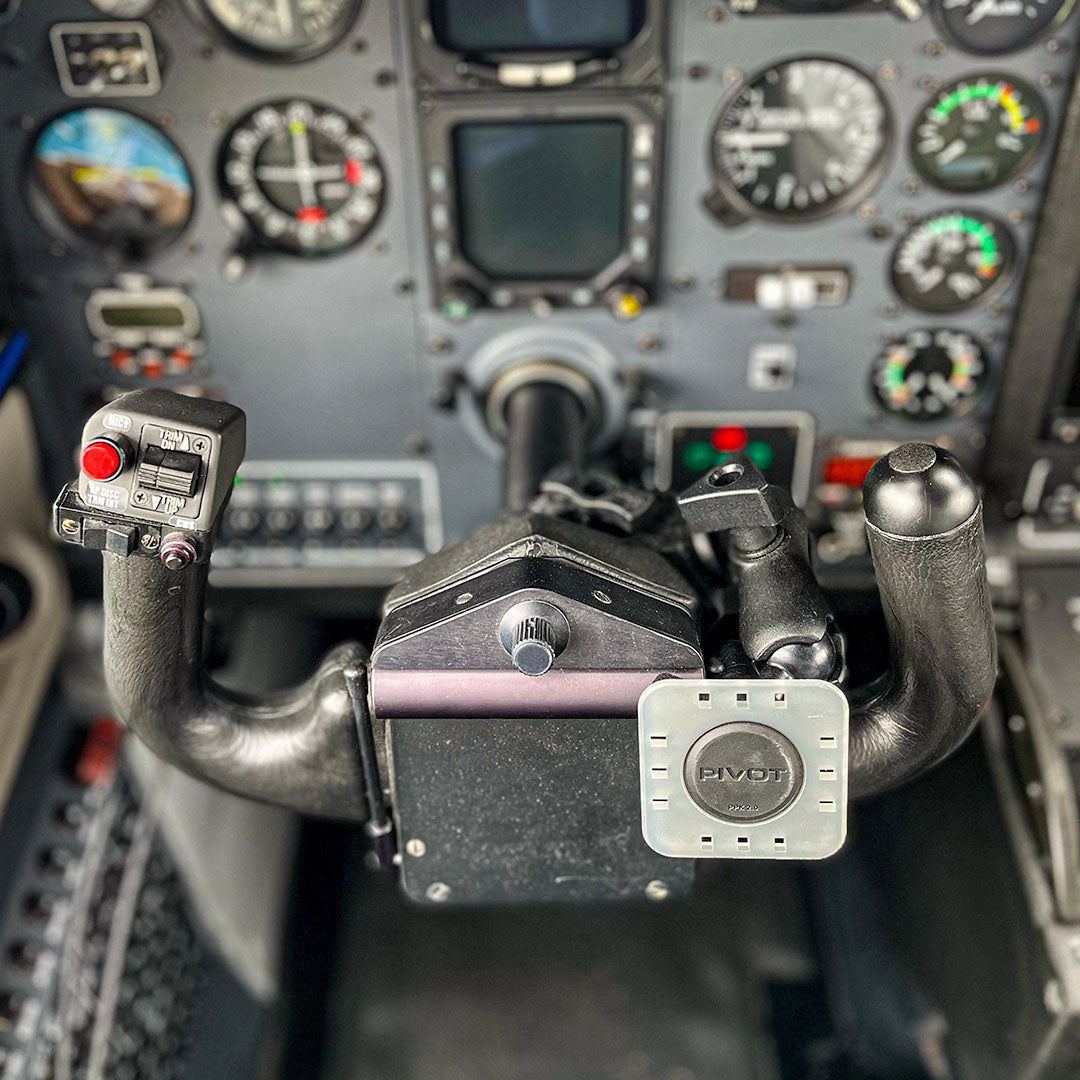 The alt text for this image is: "A close-up view of an aircraft cockpit focusing on the PIVOT Claw Mount and various flight instruments on the control panel. Buttons, dials, and a display screen are visible, indicating a complex control system.