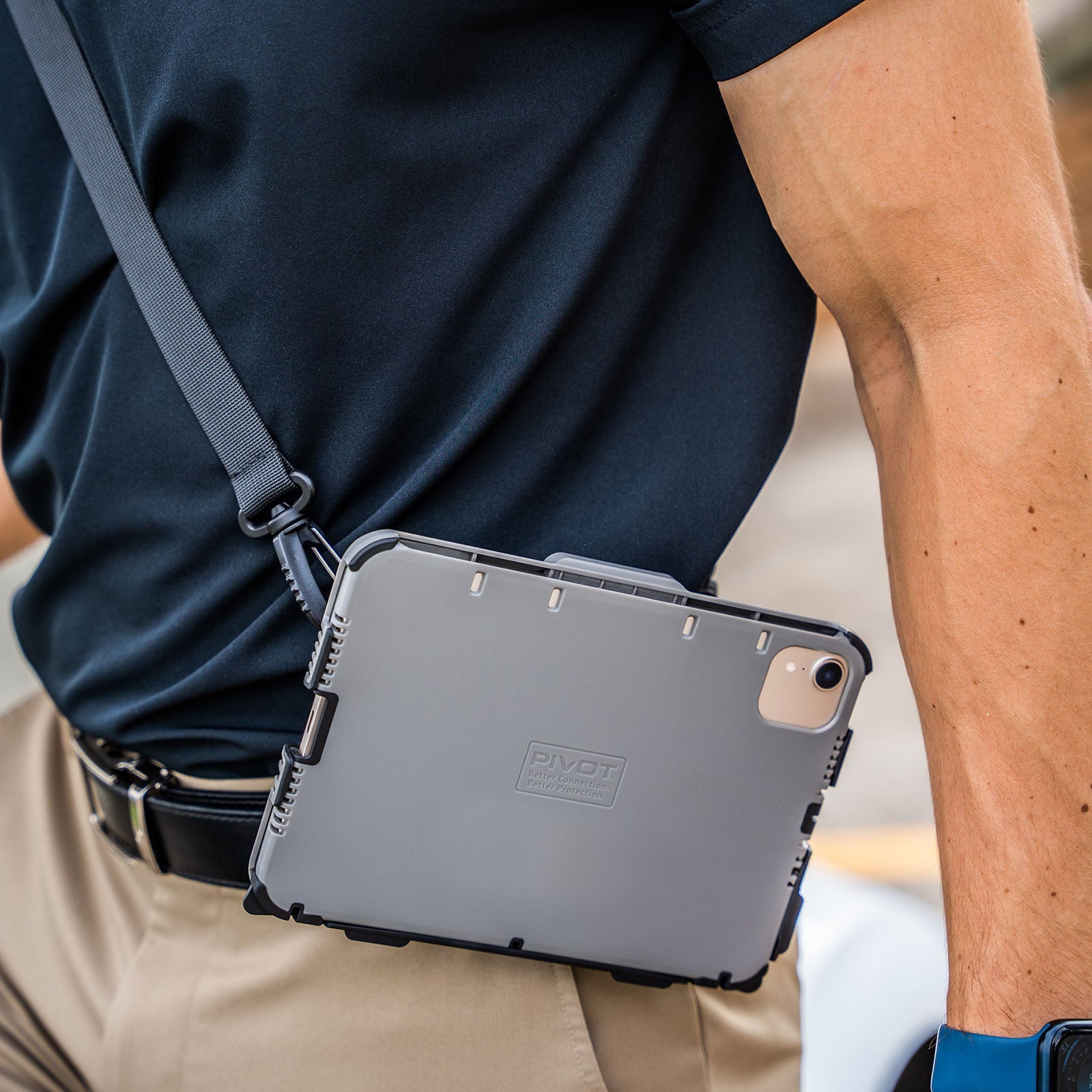 A person wearing a black shirt and khaki pants carries a PIVOT Shoulder Strap, likely attached to a tablet in a rugged case branded "PIVOT", slung over their shoulder, and they are holding a blue object in one hand.